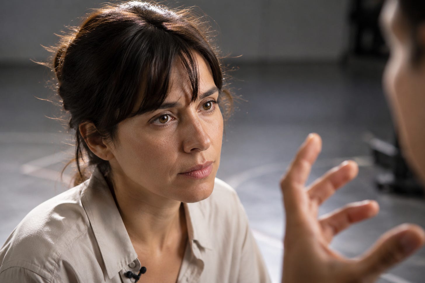 An actor's face in close-up between takes, eyes focused just off-lens, listening to an off-camera director whose hand is visible gesturing in the foreground