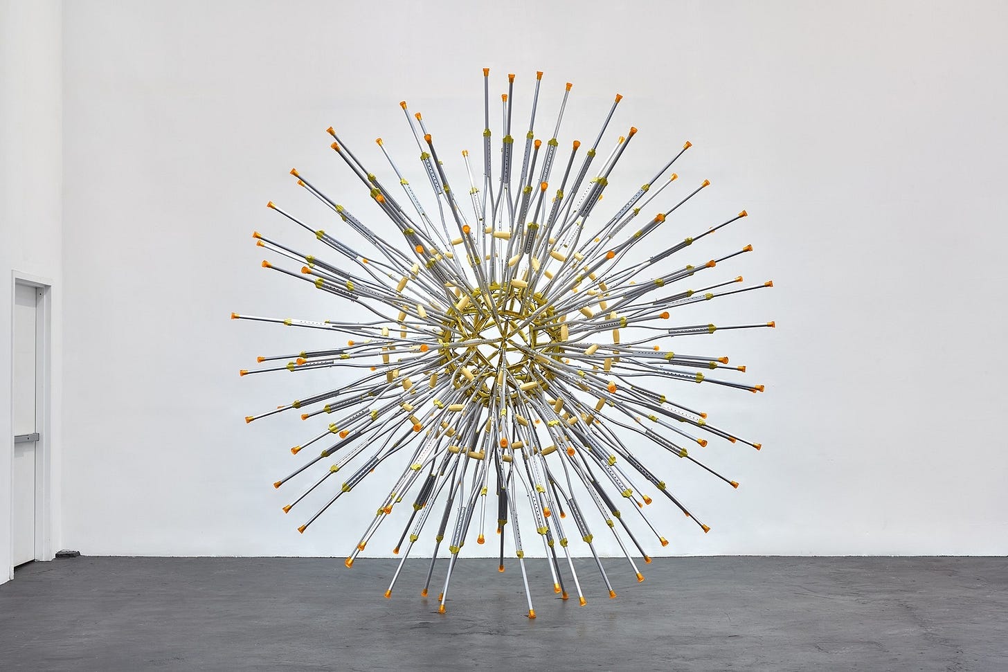 A photo of an artwork in a white walled gallery. It is a sculpture made of aluminum crutches connected to a central sphere, which looks like a huge virus, possibly Covid-19.