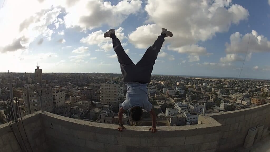 A still from the documentary Yalla Parkour — a person does a handstand on the edge of a building’s roof. A still from the documentary Yalla Parkour — a person does a handstand on the edge of a building’s roof.