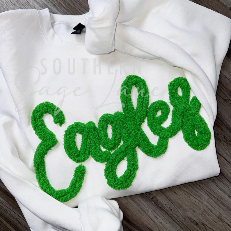 May include: White crewneck sweatshirt with a large, green, fluffy, embroidered word 'Eagles'. The embroidery is in a script font.  The sweatshirt is folded and displayed on a dark brown surface.  The brand name 'Southern Sage Lace' is subtly printed on the sweatshirt.