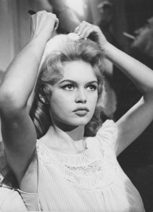 A black and white behind-the-scenes photograph of a young Brigitte Bardot styling her hair into a voluminous 1960s beehive. She is wearing a lace-trimmed white bodice and her signature winged black eyeliner and matte lip.