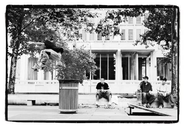 A high-contrast black-and-white photo of a skateboarder jumping off a short ramp over an outdoor garbage can while three other young skaters sit on a nearby curb watching on.