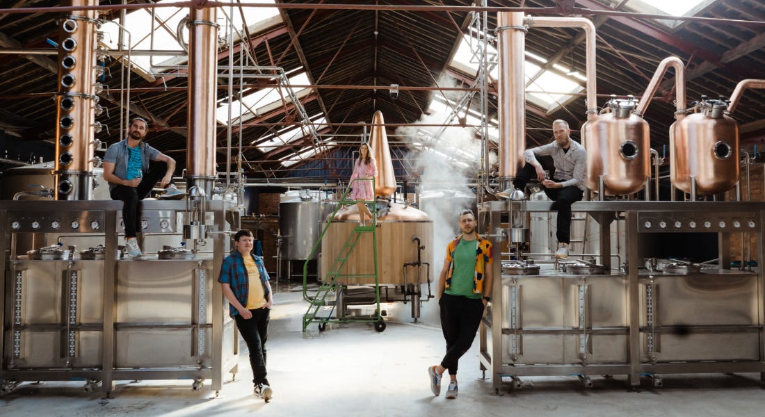 A group of five people standing around a copper rum distillery inside a barn building A group of five people standing around a copper rum distillery inside a barn building
