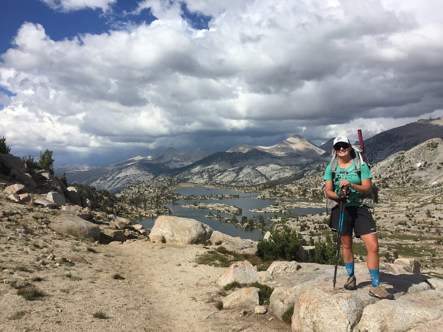 Meghan Robins hiking in granite mountains with alpine lakes and storm clouds in the distance