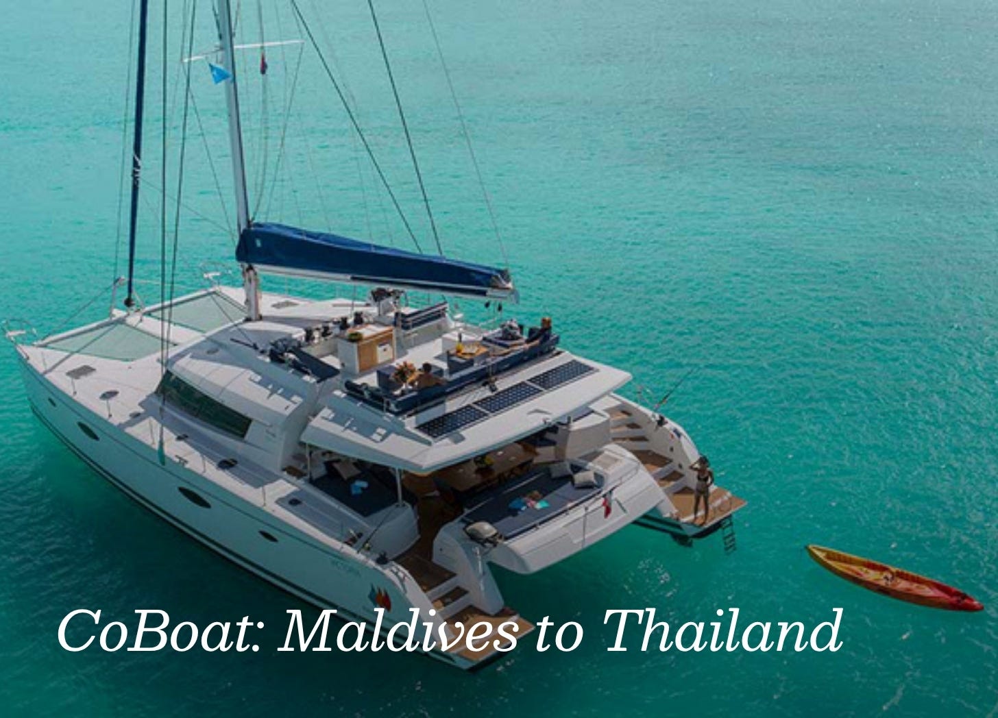Large white catamaran sits in blue-green waters, with a red kayak tethered to it and a woman added to the back deck. 