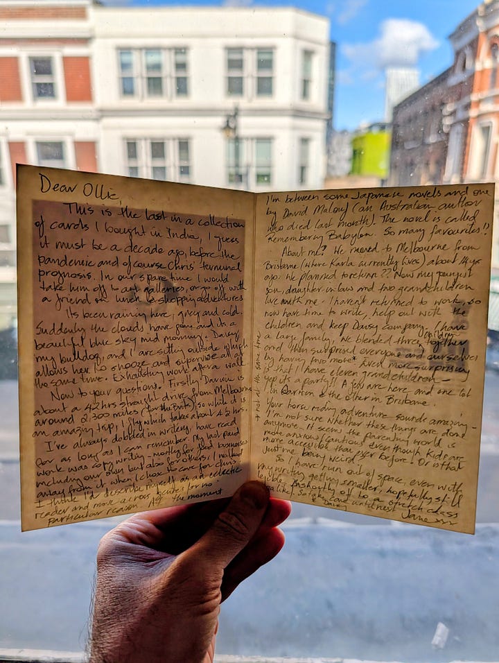 A sepia-toned watercolour and pencil portrait of a seated man wrapped in a shawl, wearing a hat. A hand holds open pages of a handwritten letter in blue ink, with a London street of brick buildings visible through a window behind.