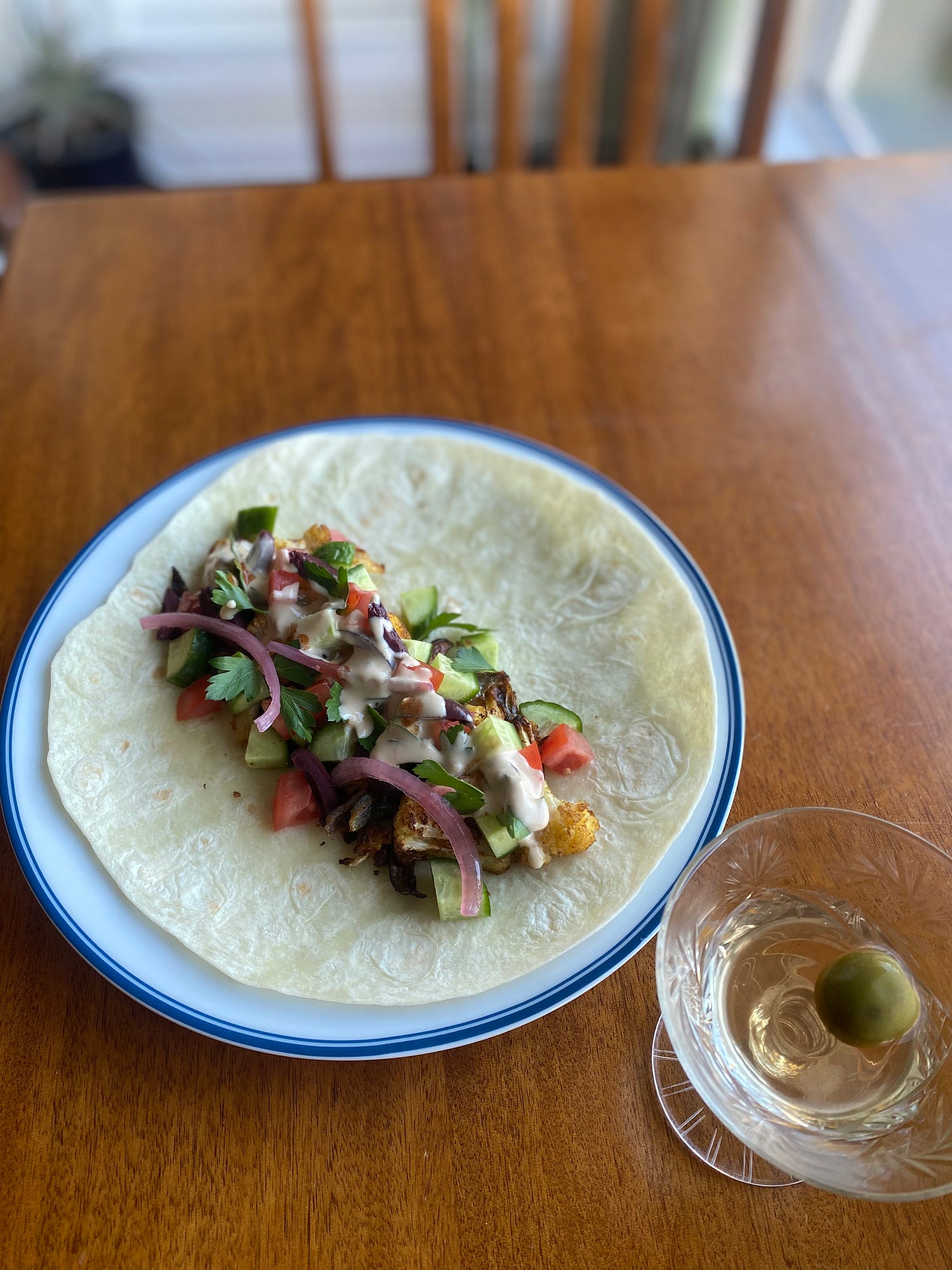 The wrap described above, fillings neatly and colourfully arranged in the centre of a tortilla on a white plate with a blue rim. The remains of a dirty martini with a castelvetrano olive is in a coupé glass next to the plate.