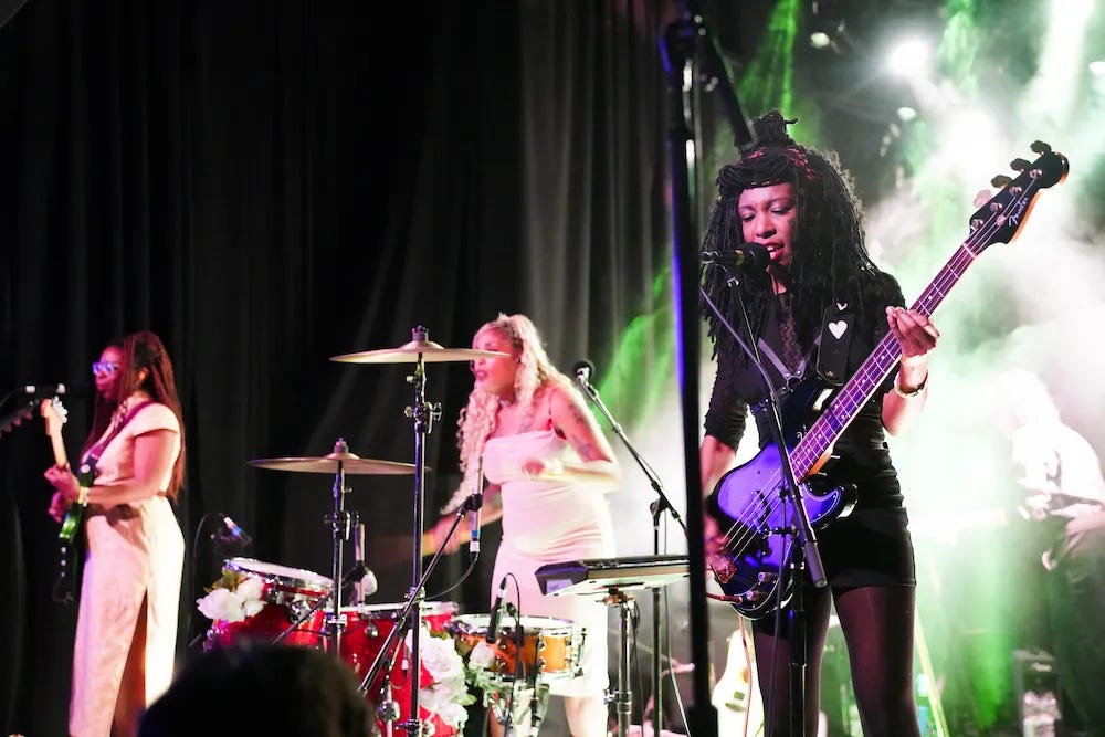 On a stage, three Black women stand in a line playing their instruments while standing at microphones. Far left is the guitarist in a white dress. In the middle, the drummer stands at her kit in a white dress. Closest to the camera is the bassist wearing all black and signing into her microphone.