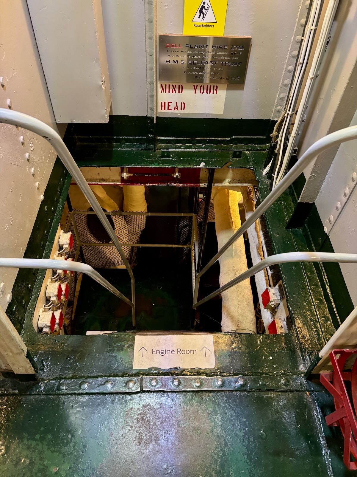 Steep metal ladder leading down to the engine room of HMS Belfast, with dim lighting and industrial surroundings.
