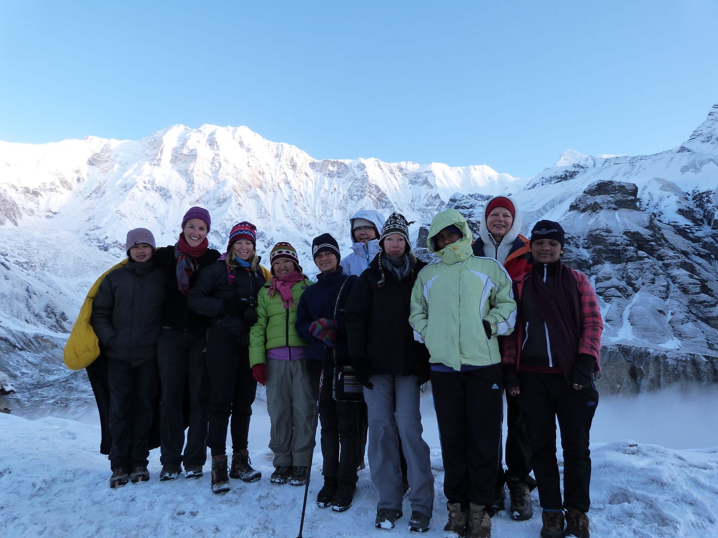 10 women wearing climbing gear standing in the snow at dawn with a snow-covered mountains towering above them.