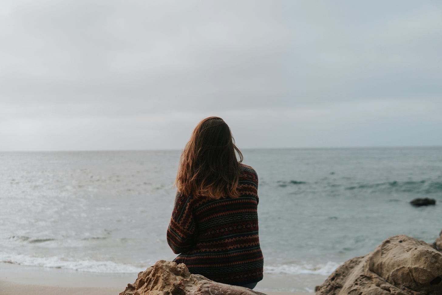 Woman with shoulder-length auburn hair sits on rock looking out at ocean, her back to the camera, it's a grey slightly-gloomy day