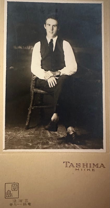 A formal portrait of a young man seated in a chair, legs crossed, in a black vest and pants with white shirt sleeves, clean shaven, looking at the camera awkwardly and yet confidently. There is Japanese and Roman text at the bottom, naming the studio.