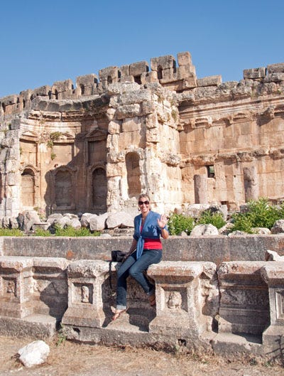 A smiling blond woman wearing jeans is sitting on an anceint wall at an archeological site. A smiling blond woman wearing jeans is sitting on an anceint wall at an archeological site.