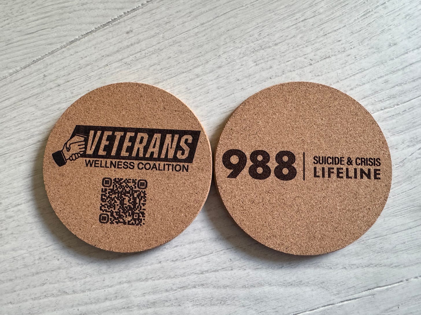 The image shows two round cork coasters on a light-colored wooden surface. One coaster displays the text "988 SUICIDE & CRISIS LIFELINE" in bold black print. The other coaster features a logo with a hand holding a cup, the words "VETERANS WELLNESS COALITION," and a QR code. The image shows two round cork coasters on a light-colored wooden surface. One coaster displays the text "988 SUICIDE & CRISIS LIFELINE" in bold black print. The other coaster features a logo with a hand holding a cup, the words "VETERANS WELLNESS COALITION," and a QR code.