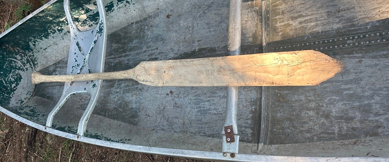 A hand-carved canoe paddle sits on top of a beat up aluminum canoe.