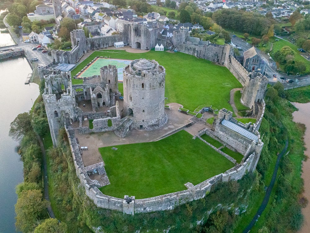 Aerial view of Pembroke Castle in Wales, showing the round Norman keep, curtain walls and inner ward, with the tidal river and town visible alongside the ruins.
