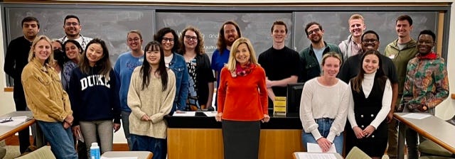 A groupd of about 25 young people stand in a classroom. A woman in a red top stands in front of the desk in the center.