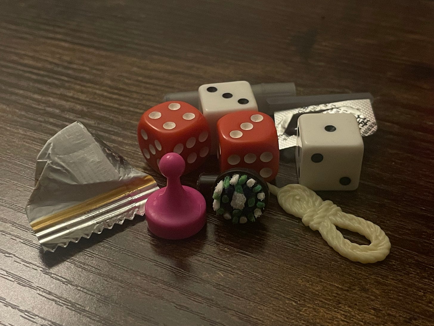 A collection of small items on a dark surface, including two red dice, two white dice, a pink pawn, a plastic token, a replacement head for an electric toothbrush, a piece of packaging for snack, and a pill in plastic packaging.
