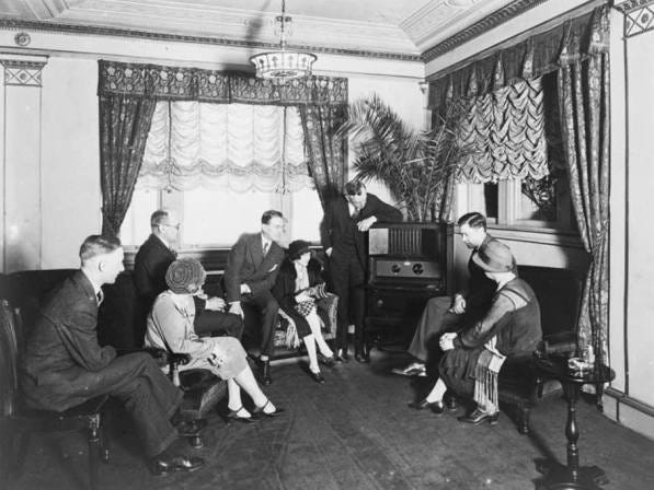 A group of people listening to the radio. The picture is in black and white and was likely taken in the 1930s. 