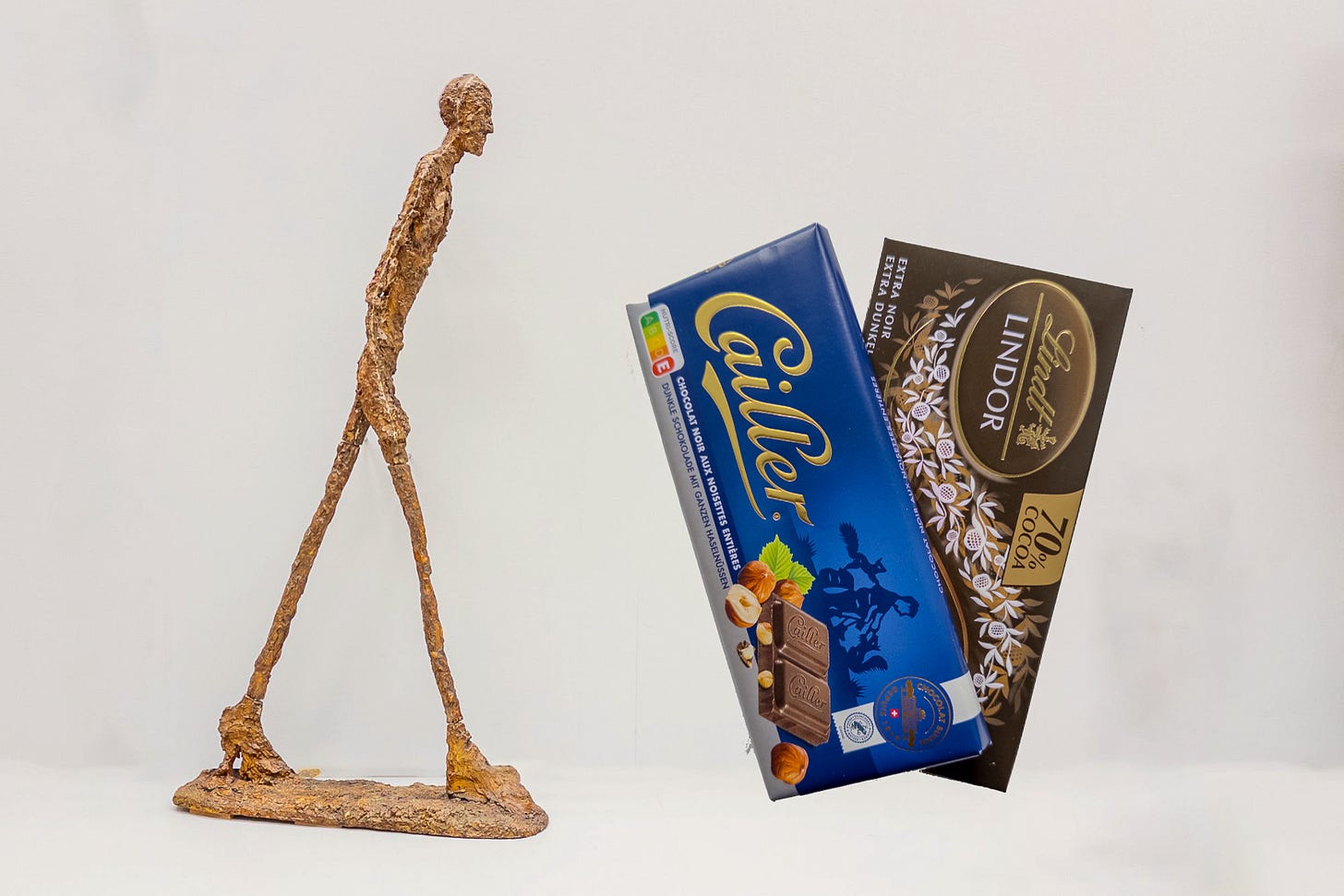 Das Bild zeigt links die überschlanke Bronzefigur „Schreitender Mann“ von Alberto Giacometti, die eine gehende Person darstellt. Rechts daneben fast gleich gross zwei Tafeln Schokolade: eine blaue Tafel der Marke Cailler mit Milchschokolade und Haselnüssen sowie eine schwarze Tafel der Marke Lindt mit dunkler Schokolade. Der Hintergrund ist hell und minimalistisch, wodurch die Objekte klar hervorgehoben werden. Das Bild zeigt links die überschlanke Bronzefigur „Schreitender Mann“ von Alberto Giacometti, die eine gehende Person darstellt. Rechts daneben fast gleich gross zwei Tafeln Schokolade: eine blaue Tafel der Marke Cailler mit Milchschokolade und Haselnüssen sowie eine schwarze Tafel der Marke Lindt mit dunkler Schokolade. Der Hintergrund ist hell und minimalistisch, wodurch die Objekte klar hervorgehoben werden.