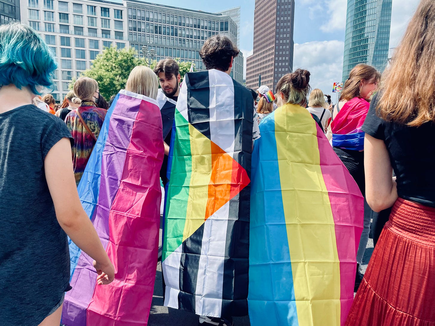 A group of people wrapped in Pride flags, including the bisexual, nonbinary, and pansexual flags, stand together in a lively crowd at Berlin CSD 2023. Tall buildings and trees are visible in the background. A group of people wrapped in Pride flags, including the bisexual, nonbinary, and pansexual flags, stand together in a lively crowd at Berlin CSD 2023. Tall buildings and trees are visible in the background.