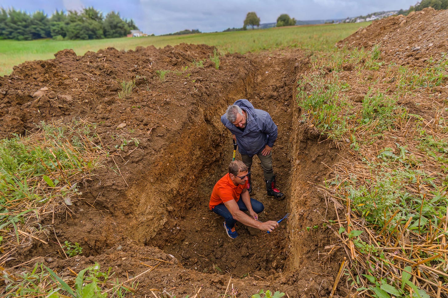 Zwei regenerative Landwirte stehen in einer tiefen Bodengrube auf einem Acker. Einer in oranger Jacke kniet und untersucht mit einem Werkzeug das Bodenprofil an der Grubenwand, der andere in grauer Jacke schaut interessiert zu. Rundherum liegen Erdhaufen und Ackerreste, im Hintergrund sind Wiesen, Bäume und ein trüber Himmel zu sehen.