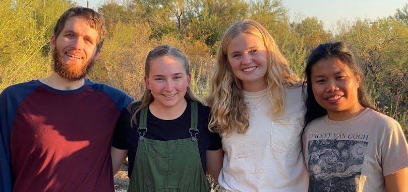 Four young adults pose together in the Sonoran Desert in Tucson. From left are a white man with a beard in a dark red shirt, a white woman wearing green overalls, a white woman wearing a white shirt and an Asian woman of color wearing a dark grey shirt.