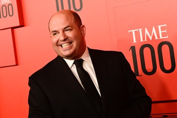 Brian Stelter, wearing a jacket and tie, smiles in front of a red backdrop bearing the logo for Time magazine’s list of the 100 most influential people.