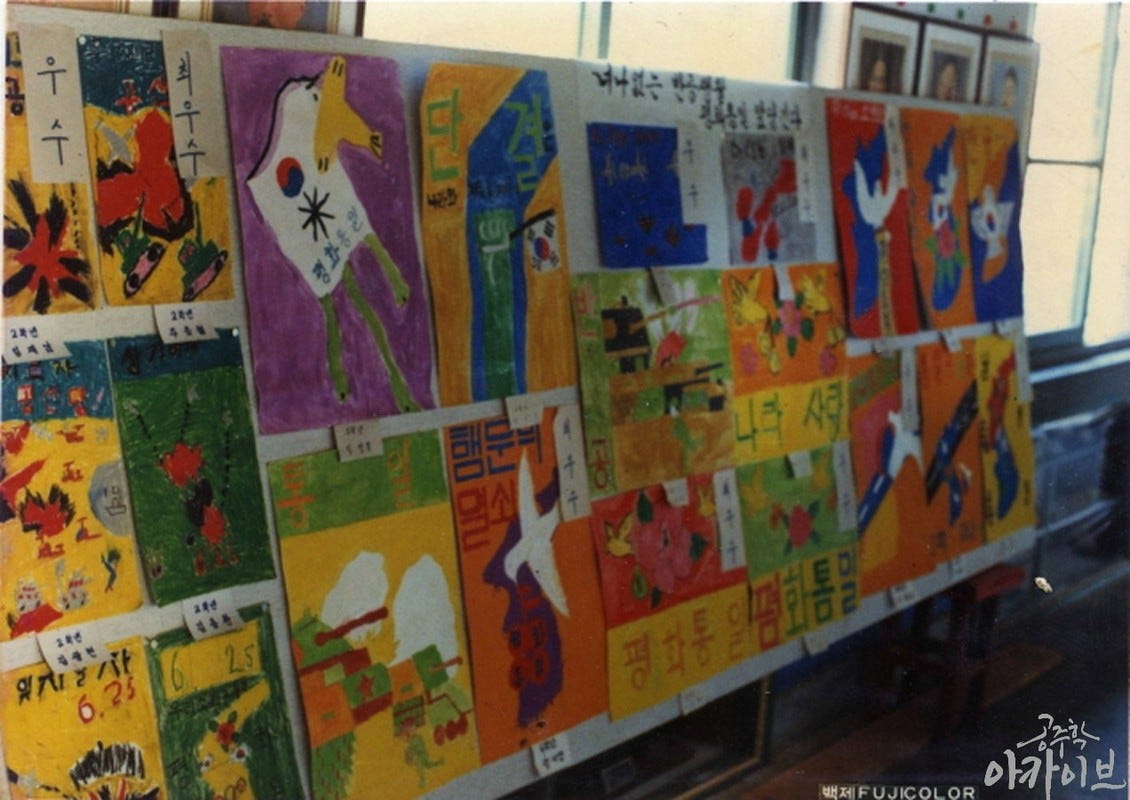A classroom display board filled with colorful anti-communist posters made by elementary school students in 1987. The posters feature doves, tanks, the South Korean flag, slogans like "Unification through Peace," and visual references to the Korean War. Each poster is labeled with handwritten titles and award tags.