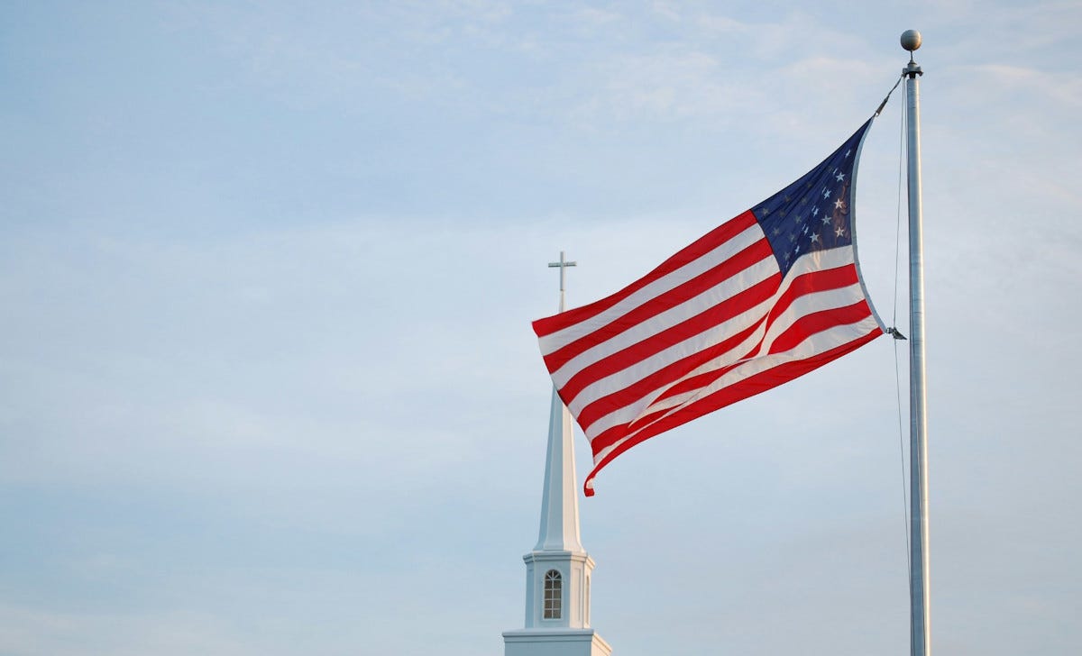 Steeple with a cross on top and a U.S. flag flying in the foreground Steeple with a cross on top and a U.S. flag flying in the foreground