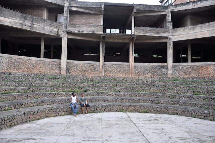 A man and a woman sit on stairs made from stone in an arena with a concrete building behind them.