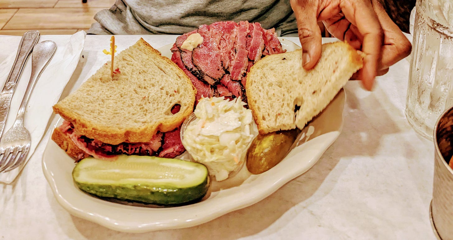 Pastrami sandwich, with one side open faced with a dab of Russian dressing. The plate also has pickles and coleslaw.