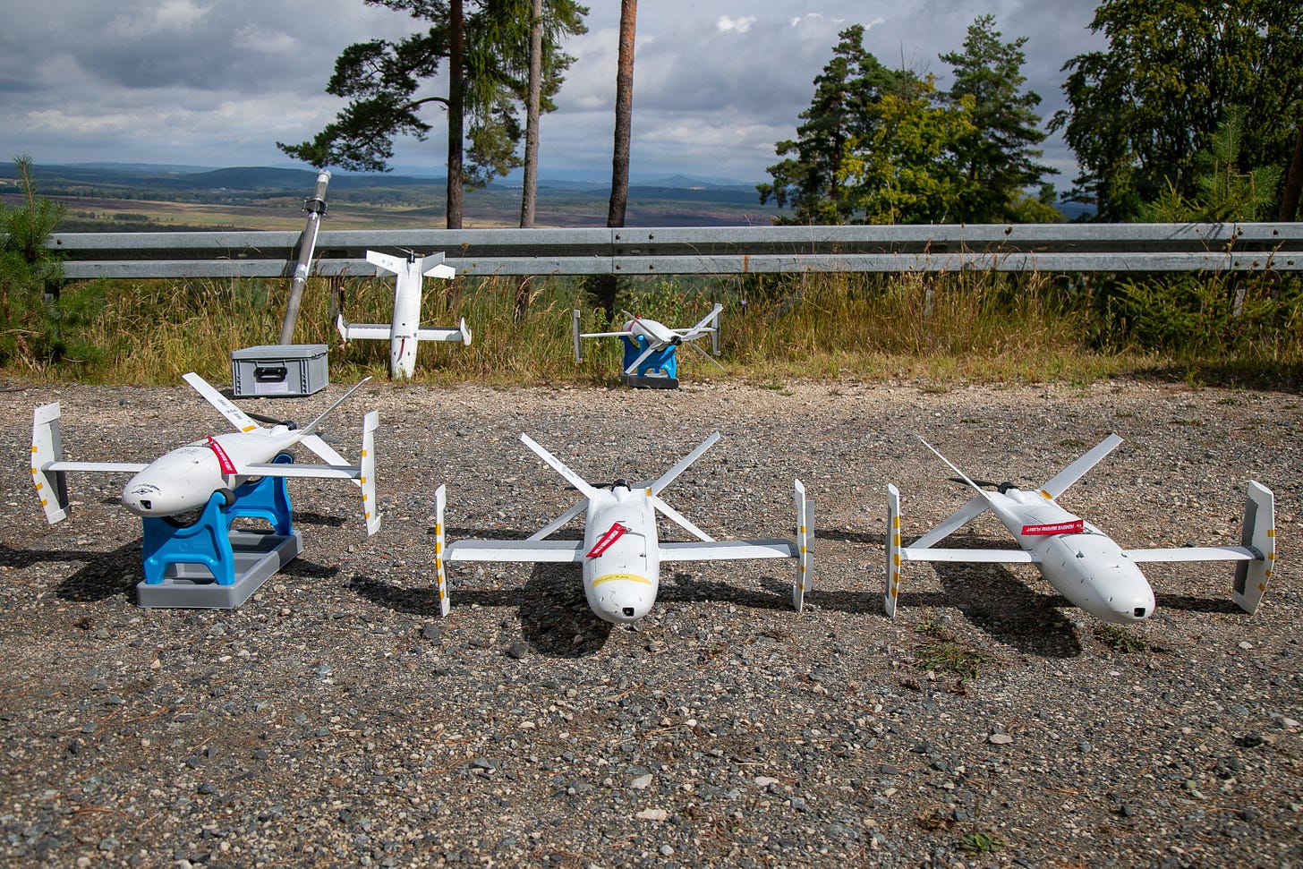 Four white fixed-wing target drones parked on gravel at a training area, with forest and hills behind