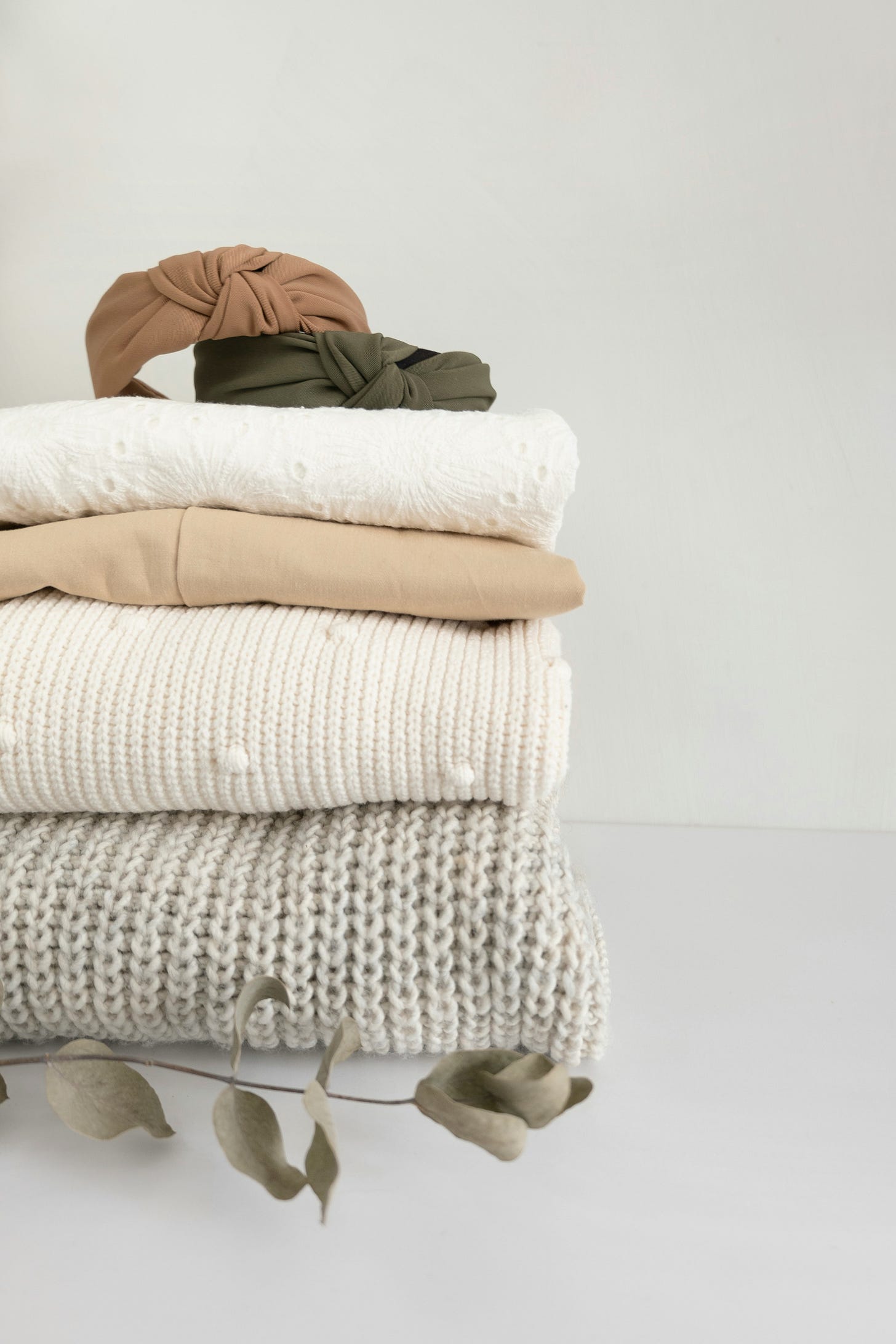 A stack of neutral-toned blankets and clothing, topped with a brown and an olive green knotted headband. Eucalyptus leaves lie nearby, creating a serene, cozy atmosphere. A stack of neutral-toned blankets and clothing, topped with a brown and an olive green knotted headband. Eucalyptus leaves lie nearby, creating a serene, cozy atmosphere.