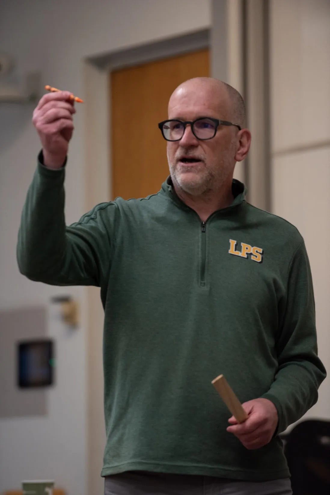 A man wearing glasses and a green quarter-zip pullover with the letters "LPS" on the chest gestures with his right hand while holding a small orange object, possibly a pen or pencil. In his left hand, he holds a rectangular wooden stick. He appears to be speaking or presenting in an indoor setting, possibly a classroom or lecture hall.