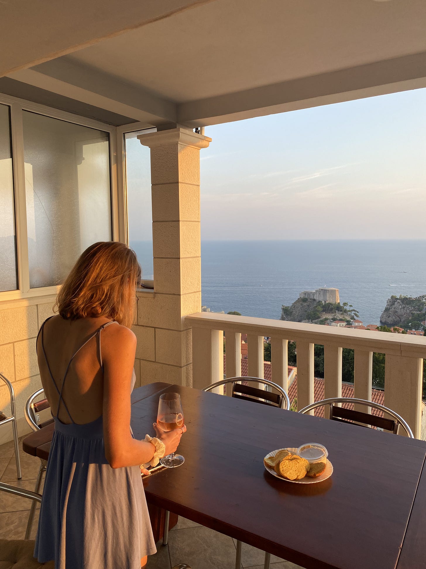 A young blonde woman holding a glass of wine and looking out to the sea and sunset from a balcony, with a brown wooden table on which there is some bread and hummus on a plate.
