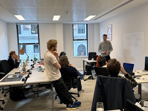 Four men smiling at the camera with a red computer chip on the table in front of them (left hand side), three men sat down on chairs while one speaks in front of a whiteboard (right hand side)