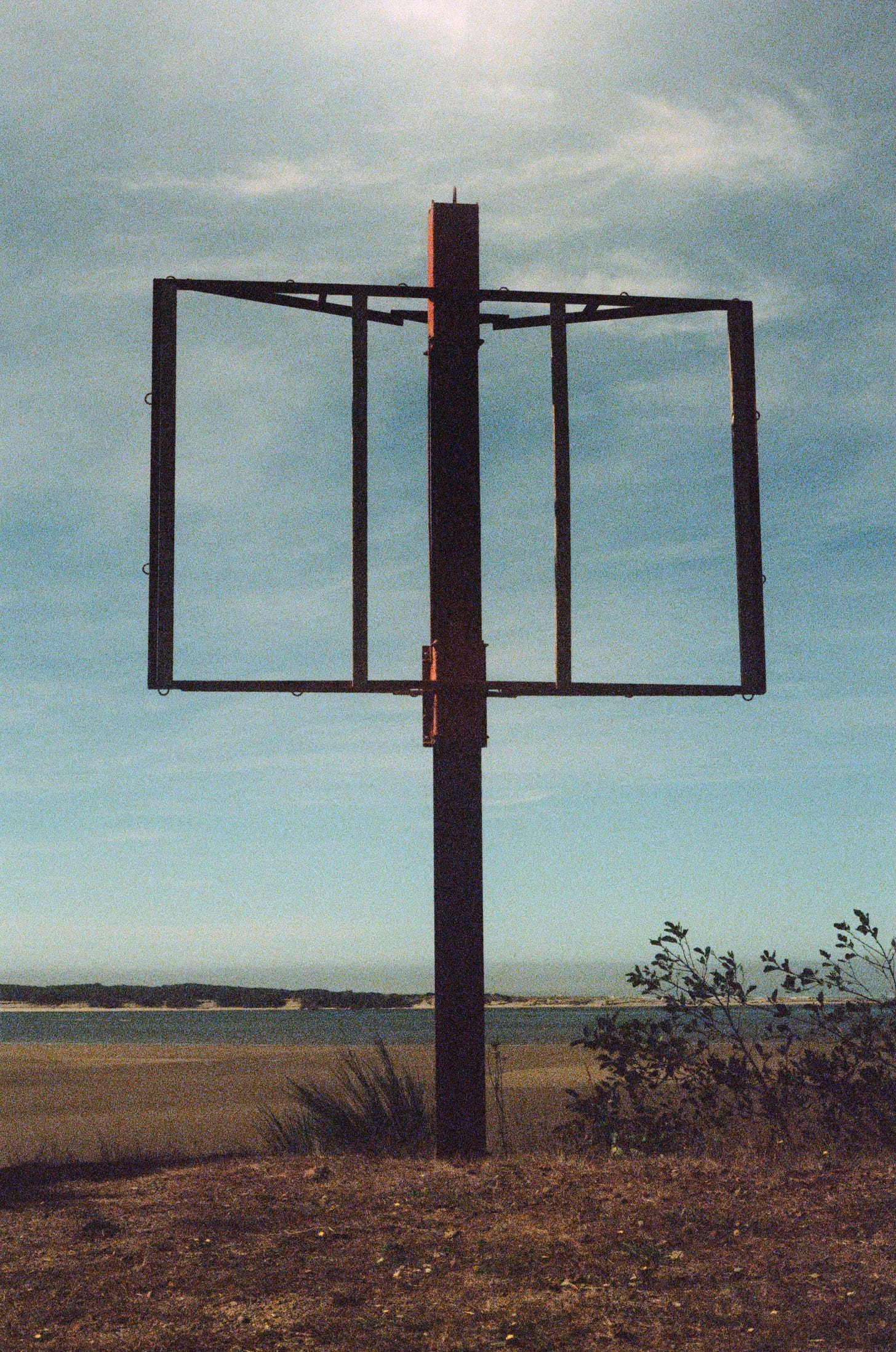 A slightly grainy film photograph of a large metal frame that seems to have once held a billboard. It stands against a blue sky with minimal clouds, which can also be seen through the frame itself.