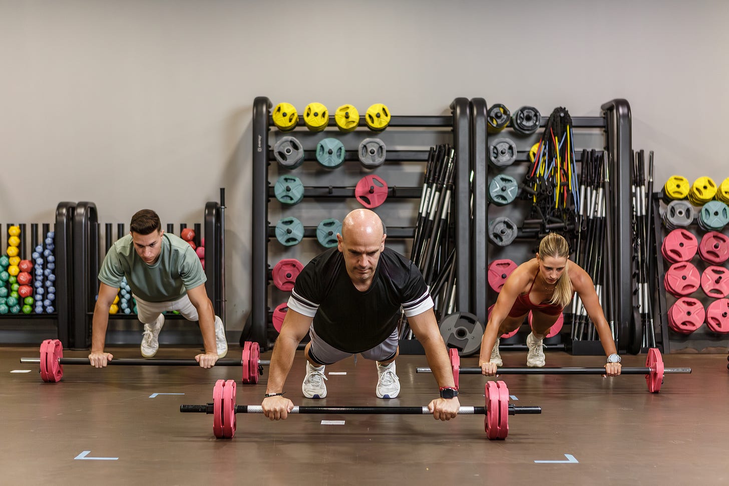 Three people in a group fitness room at the gym, doing planks with hands on dumbbells. Three people in a group fitness room at the gym, doing planks with hands on dumbbells.