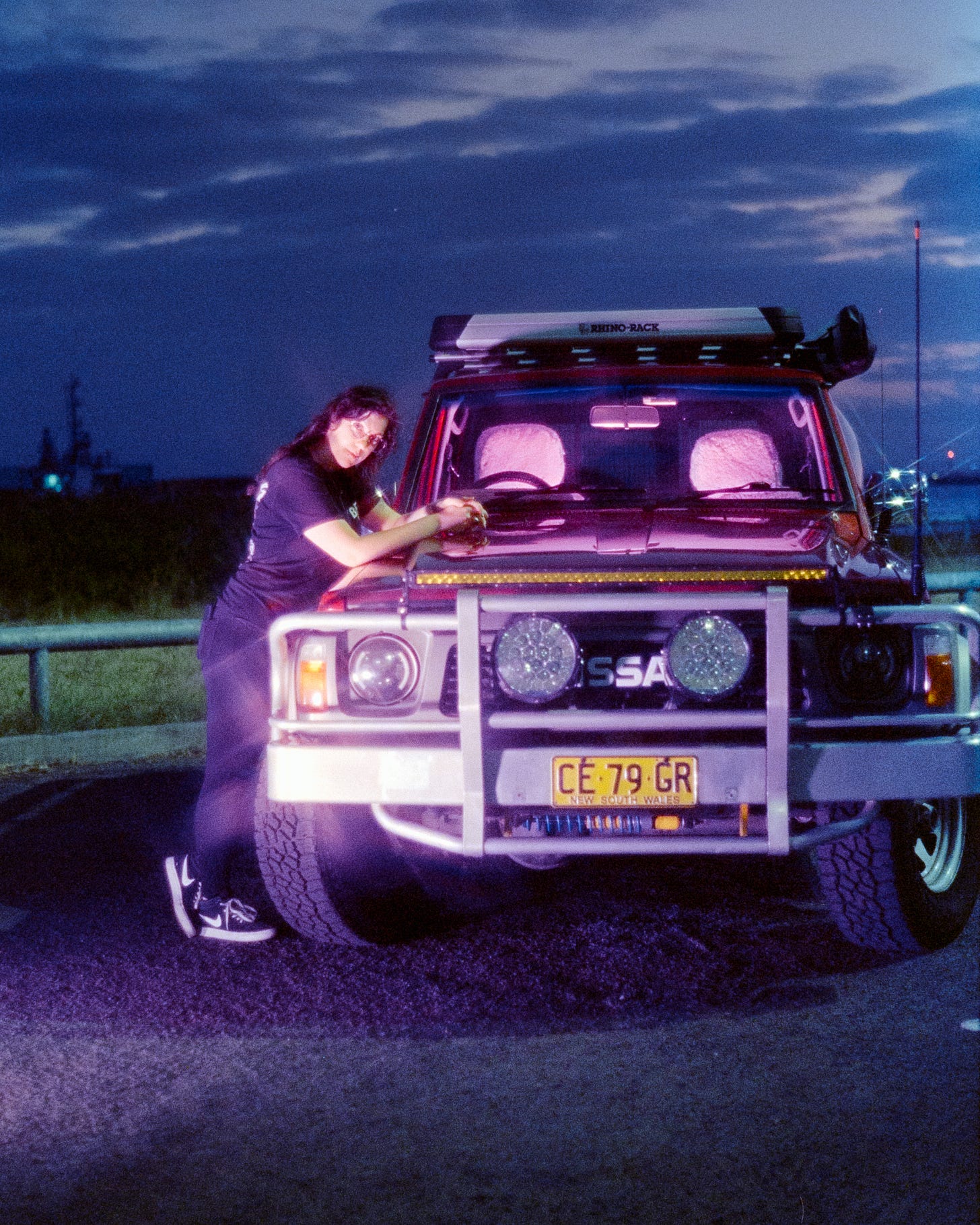 Josie standing to the side of her large 4WD, lovingly named Scorpion. Both are illuminated by bright purple lights. Josie is leaning against the hood with hands collapsed, smiling at the camera. Josie standing to the side of her large 4WD, lovingly named Scorpion. Both are illuminated by bright purple lights. Josie is leaning against the hood with hands collapsed, smiling at the camera.