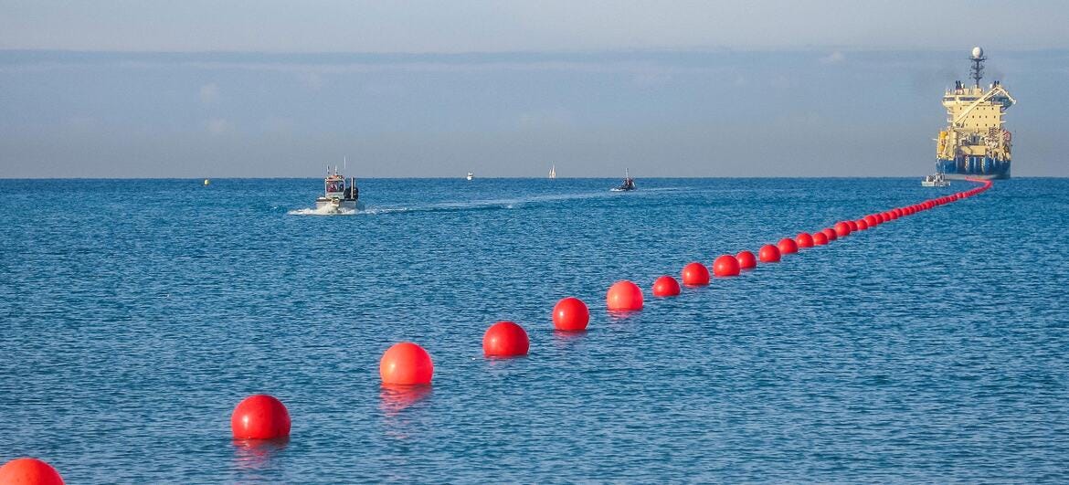 A line of red buoy markers in the ocean, with a cable laying vessel and small boats in the distance, representing the International Submarine Cable Resilience Summit 2026.