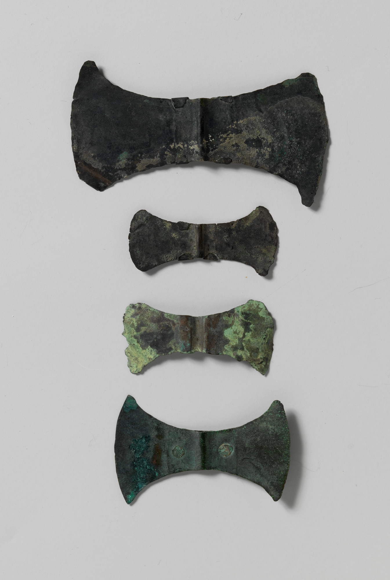 Four heavily worn and corroded bronze labrys blades of varying sizes, without handles, are arranged in a vertical row on a white background.