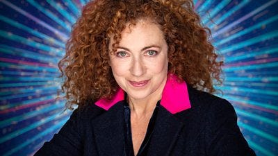 Head and shoulders shot of Alex Kingston, a woman with curly hair wearing a dark jacket with bright pink lapels. She smiles to camera.