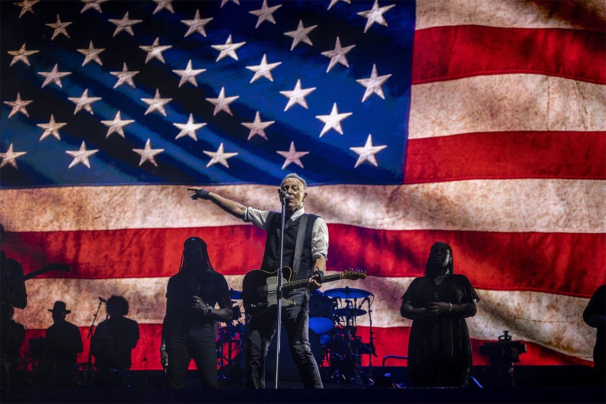 Bruce Springsteen performs with the E Street Band during a concert in Washington, D.C., with musicians playing instruments and engaging the audience with the United States flag as a backdrop.