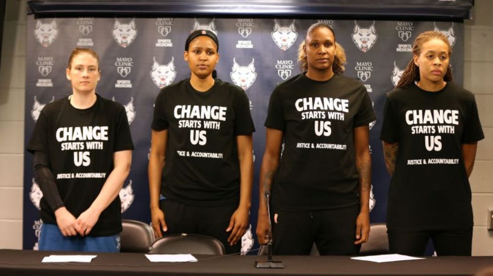 Four Minnesota Lynx players stand in front of a press conference table wearing black shirts that read "Change Starts With Us: Justice.& Accountability"