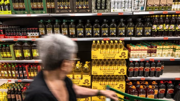 Donna con capelli bianchi in piedi davanti alla sezione degli oli di un supermercato, con scaffali pieni di bottiglie e prodotti disposti ordinatamente. L'ambiente è ben illuminato e moderno. Donna con capelli bianchi in piedi davanti alla sezione degli oli di un supermercato, con scaffali pieni di bottiglie e prodotti disposti ordinatamente. L'ambiente è ben illuminato e moderno.