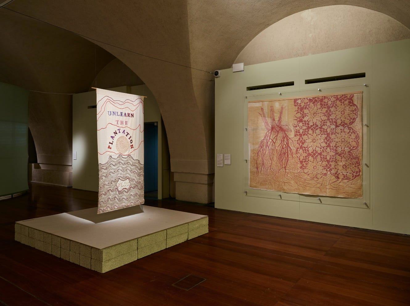 A tapestry artwork embroidered with the slogan "UNLEARN THE PLANTATION" in an eerily empty gallery space.