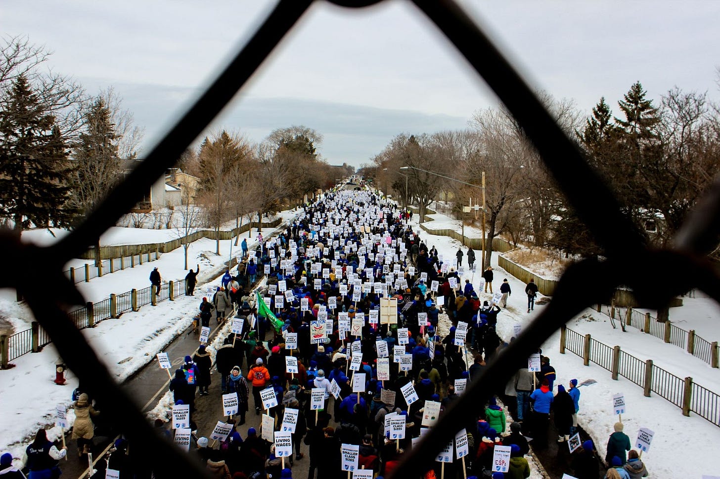 an aerial view through a chain link fence of a crowd of protesters marching through the streets carrying picket signs. snow lines the sidewalks and many protesters wear blue hats