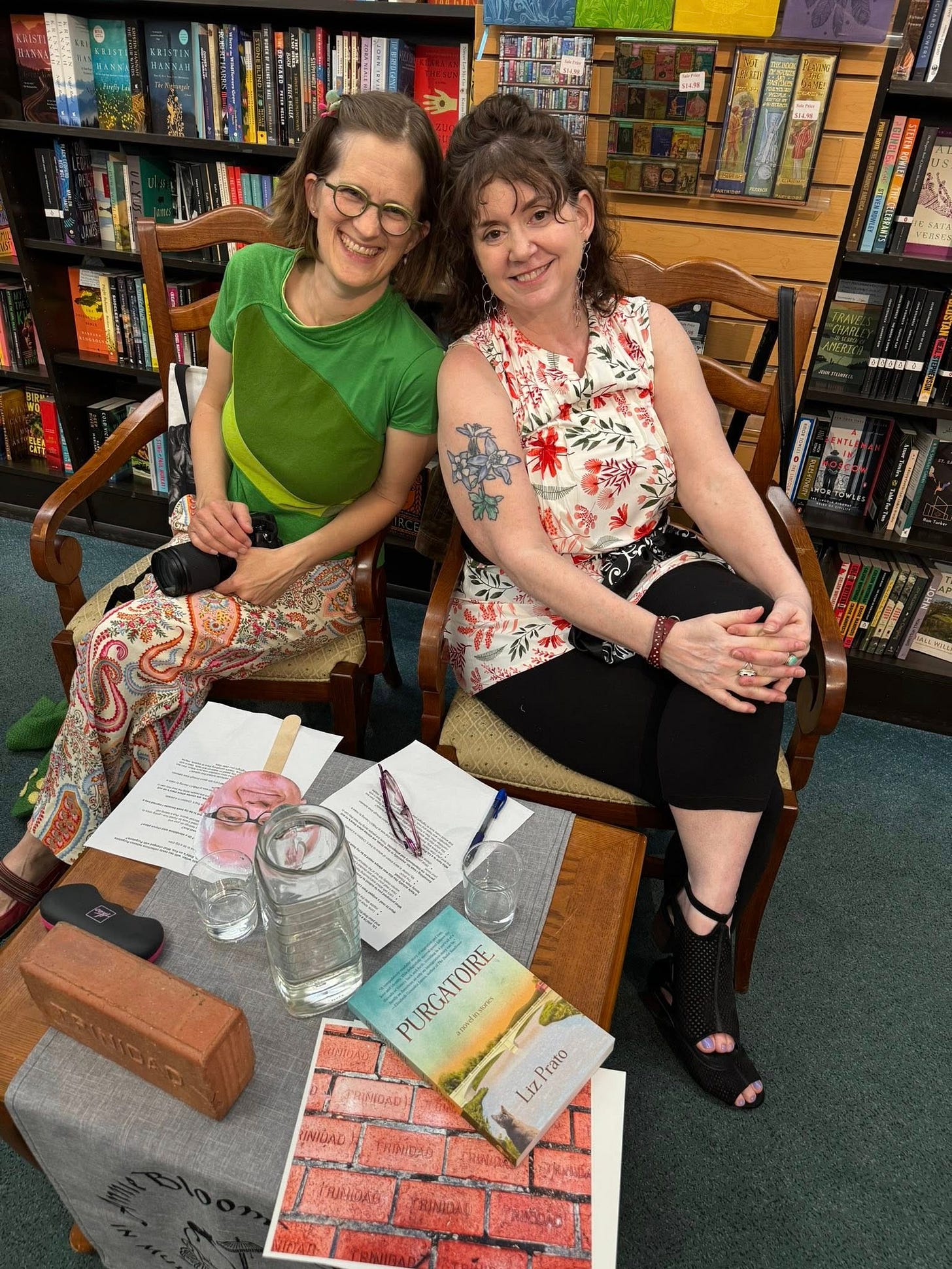 Two white women lean against each other in chairs with a bookstore background. Two white women lean against each other in chairs with a bookstore background.