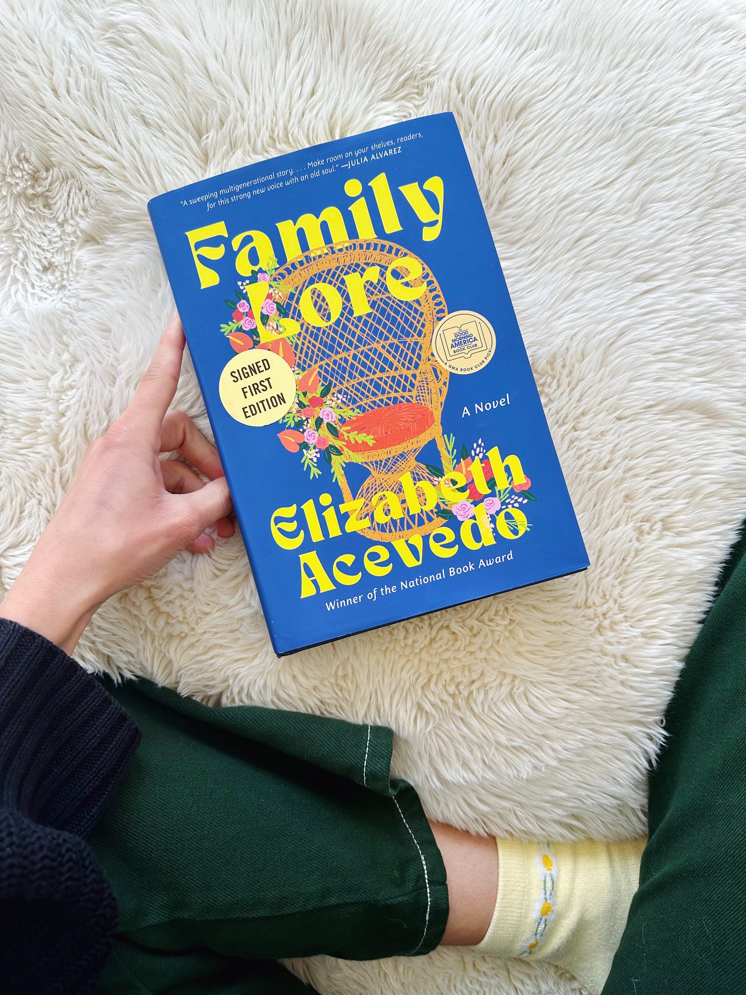 A white hand holds Family Lore against a fluffy, white blanket